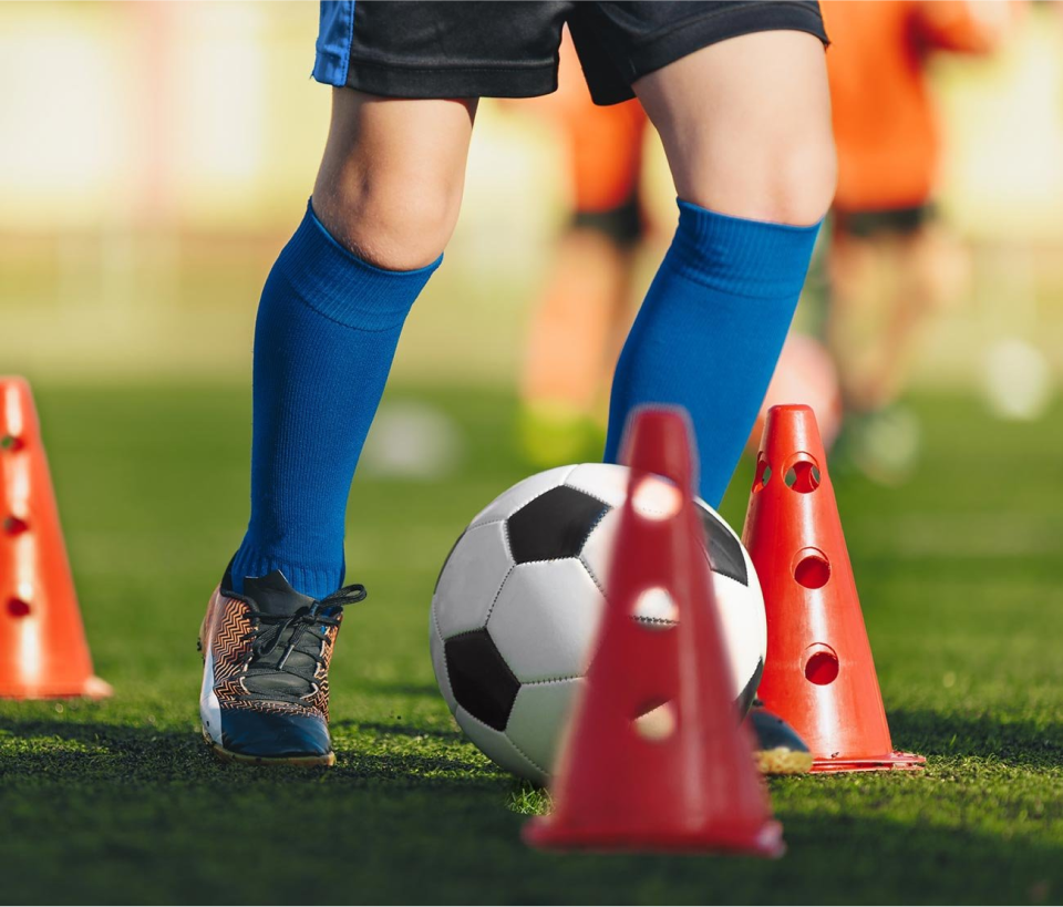 Image of a young kid playing training soccer with obstacles in a grass field, wearing blue socks, black shorts with blue stripes on the side.